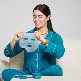Model with The Skin Nerd Hydra-Hit Instant Facial Mask, holding up blue sheet mask, seated in teal pyjamas with gold necklace