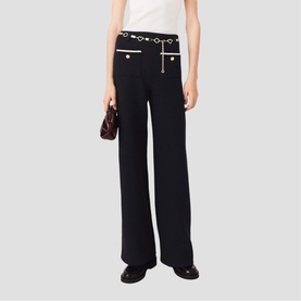 Model wearing Maje wide-leg trousers in black with gold-tone heart hardware belt detail, styled with a brown bag and black shoes.