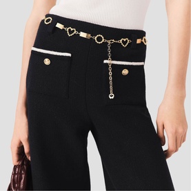 Model wearing Maje black wide-leg trousers with gold heart chain belt and contrast white trim pockets, styled with burgundy bag