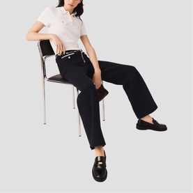 Model carrying Maje red bag, styled with white polo shirt, black wide-leg trousers and black loafers, seated on chrome chair