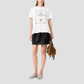 Model wearing Maje white graphic T-shirt and black mini skirt, styled with metallic flats and carrying a tan fringed bag.
