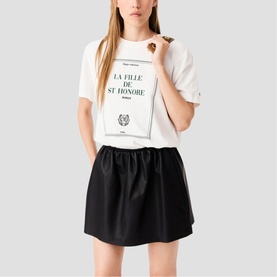 Model wearing Maje white graphic T-shirt with "La Fille de St Honor&eacute;" print, styled with a black mini skirt.