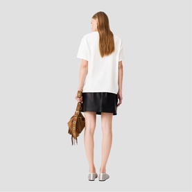 Model carrying Maje bag in metallic tan, with fringe and stud detailing, styled with white oversized tee and black shorts