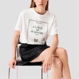 Model wearing Maje white graphic T-shirt with La Fille de St Honor&eacute; print, styled with a black mini skirt, seated on a chair
