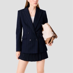 Model wearing Maje navy pinstripe double-breasted blazer and pleated mini skirt, styled with a beige tote bag.