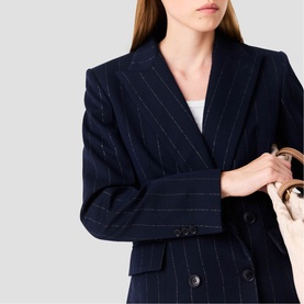 Model wearing Maje navy pinstripe double-breasted blazer, styled with a white top and carried woven bag visible at side.