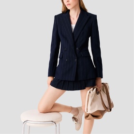 Model wearing Maje Womens French Dress in navy pinstripe, styled with a beige pleated mini skirt, carrying a tan structured bag with nude ankle boots.