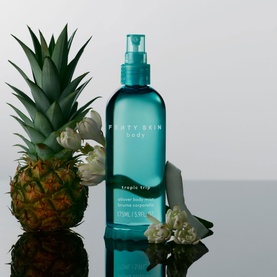 Fenty Skin Body Tropic Trip Shimmer Body Mist in teal spray bottle, styled with pineapple and white flowers on reflective surface