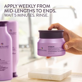 Pureology Hydrate Hair Treatment Mask in purple pot held by hand, with matching shampoo bottles visible in background