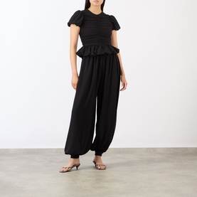 Model wearing Rotate black short-sleeve ruched peplum top, styled with black wide-leg trousers and silver strappy heels.