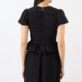 Model wearing Rotate black top, back view, showing ruched bodice, flutter sleeves, and peplum hem detail in polyester fabric.