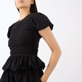 Model wearing Rotate black ruched short-sleeve top with puffed cap sleeves and peplum hem detail, paired with black tiered skirt.