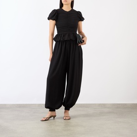 Model wearing Rotate black smocked puff-sleeve top with ruffle hem, styled with black wide-leg trousers and nude kitten heels.