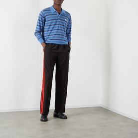 Model wearing Wales Bonner Energy Logo Track Pants in Brown/Black with red contrast leg stripe, paired with a blue striped polo and black shoes.