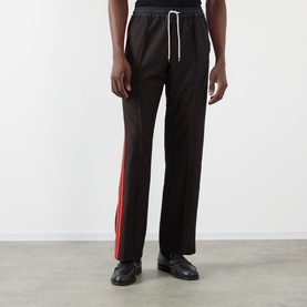 Model wearing Wales Bonner Energy Logo Track Pants in brown/black with red contrast side stripe, straight-leg fit and elasticated drawcord waist.