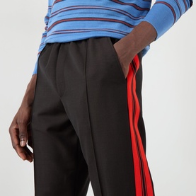 Model wearing Wales Bonner Energy Logo Track Pants in brown/black with red contrast side stripes, styled with a blue striped top.