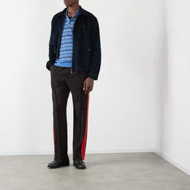 Model wearing Wales Bonner Energy Logo Track Pants in Brown/Black with red contrast leg stripe, styled with navy jacket and blue stripe polo.
