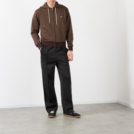 Model wearing Wales Bonner Brown/Caramel zip-up hoodie with allover print, styled with black wide-leg trousers and dark trainers.