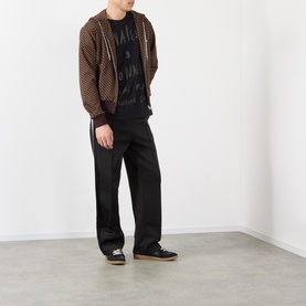 Model wearing Wales Bonner brown/caramel zip-up hoodie with black graphic tee, black wide-leg trousers and dark trainers.