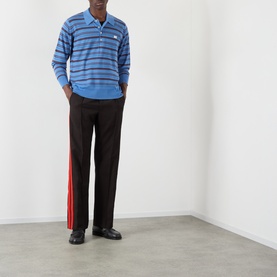 Model wearing Wales Bonner Beam Logo Striped Polo Sweater in Chambray Blue, styled with dark trousers featuring red side stripes and black loafers.