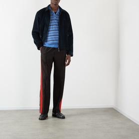 Model wearing Wales Bonner Beam Logo Striped Polo Sweater in Chambray Blue, styled with navy jacket, brown trousers with red side stripe, and black