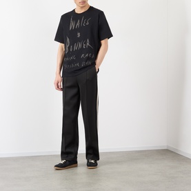 Model wearing Wales Bonner black graphic cotton T-shirt, styled with wide-leg black trousers and dark low-top trainers.