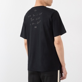 Model wearing Wales Bonner black cotton T-shirt, back view showing tonal handwritten-style text graphic, styled with black trousers.