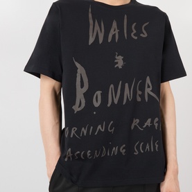 Model wearing Wales Bonner black cotton T-shirt with tonal hand-written logo and text graphic print to front.