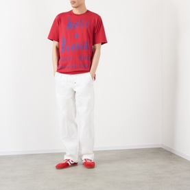 Model wearing Wales Bonner Raga Logo T-Shirt in Rooibos red with blue logo lettering, styled with white wide-leg trousers and red trainers.