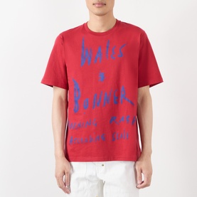 Model wearing Wales Bonner Raga Logo T-Shirt in Rooibois red with blue logo lettering, crew neck, short sleeves, styled with white trousers