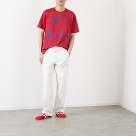 Model wearing Wales Bonner Raga Logo T-Shirt in Rooibois red with blue logo lettering, styled with white wide-leg trousers and red trainers.