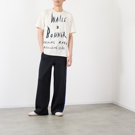 Model wearing Wales Bonner Ivory cotton T-shirt with handwritten graphic print, styled with wide-leg navy trousers and white trainers.