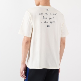 Model wearing Wales Bonner ivory cotton T-shirt, back view showing handwritten-style text graphic, paired with blue trousers.