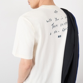 Model wearing Wales Bonner ivory cotton T-shirt, back view showing handwritten-style script print, styled with navy outerwear draped over shoulder.