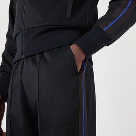 Model wearing Wales Bonner Form Logo Track Pants in black with blue and brown contrast side stripes, styled with matching zip-up track jacket.