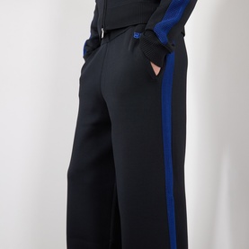 Model wearing black wide-leg trousers with blue side stripe, styled with matching black zip-up top with blue panel detail, hands in pockets