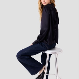 Model wearing Maje top in navy, seated on white stool, styled with dark wide-leg jeans with contrast side stripe