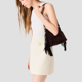 Model carrying Maje dark brown suede shoulder bag with fringe detail and gold hardware, styled with white mini dress.