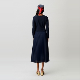 Model wearing Claude Pierlot blue pleated midi skirt and navy knit top, styled with red headscarf and gold heeled mules, back view.