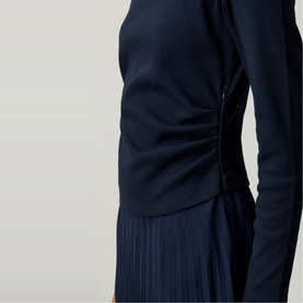 Model wearing Claude Pierlot blue long-sleeve top with ruched side detail, styled with a navy pleated skirt.