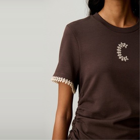 Model wearing Claude Pierlot brown T-shirt with gold embroidered C logo and daisy-chain trim on sleeves, ruched side detail.