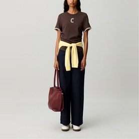 Model wearing Claude Pierlot brown logo T-shirt, styled with navy wide-leg trousers, yellow tied jacket and burgundy bucket bag.