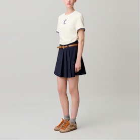 Model wearing Claude Pierlot top in clear, styled with navy pleated mini skirt, tan belt, and chunky orange trainers.