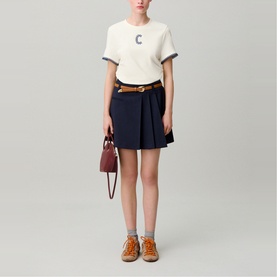 Model wearing Claude Pierlot cream top with navy pleated mini skirt and tan belt, styled with burgundy bag and grey lace-up shoes.