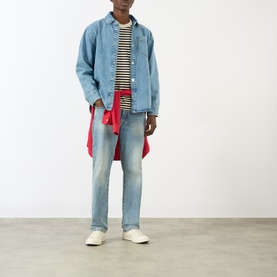 Model wearing AMI Bleu Used denim casual jacket, styled with striped tee, light wash jeans, red shirt tied at waist and white trainers.