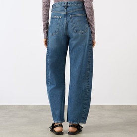Model wearing AgoldE jeans in proof blue denim, rear view, relaxed wide-leg fit, styled with a mauve knit top and black sandals.