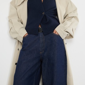 Model wearing AgoldE rinse wide-leg jeans in dark indigo denim, styled with a navy knit vest and beige trench coat, belt visible at waist.