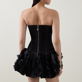 Model wearing Aje black strapless mini dress with ribbed corset bodice, gold zip back, and voluminous ruffled organza skirt.