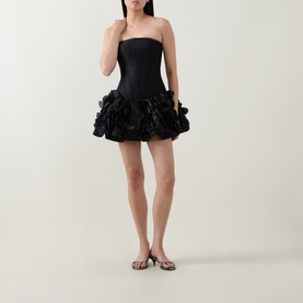 Model wearing Aje black strapless mini dress with voluminous floral ruffle skirt, styled with brown flat sandals