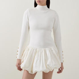 Model wearing Aje cream dress with ribbed high-neck long-sleeve bodice, button cuff detail, and voluminous bubble skirt hem.